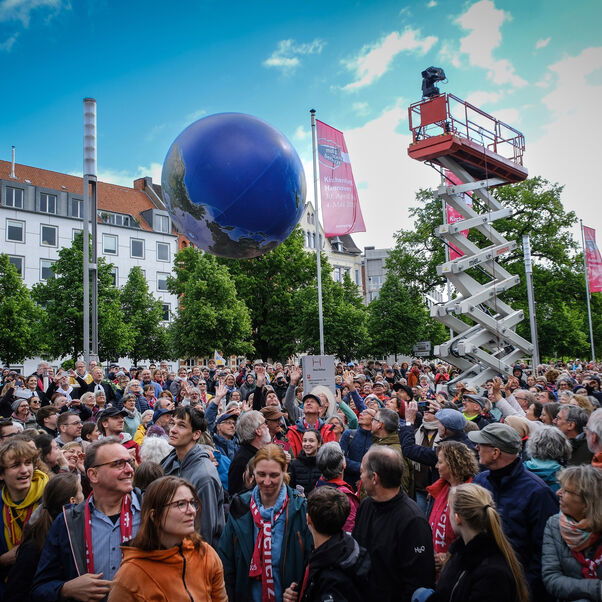 Eine Menschenmenge schaut auf einem sehr großen Ballon, der wie die Erde gestaltet ist, und von den Menschen mit den Händen weitergeleitet wird.