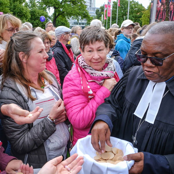 Eine männlich zu lesende Person teilt Oblaten aus in einem Gottesdienst unter freiem Himmel.