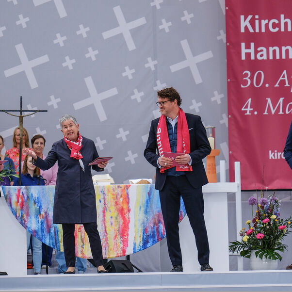 Drei männlich und eine weiblich zu lesende Personen, alle in schwarz gekleidet mit roten Schals, stehen auf einer Bühne.