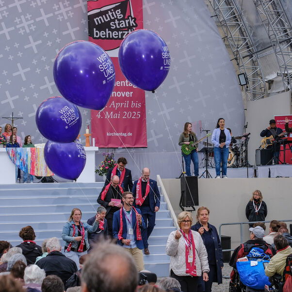 Personen gehen auf einer Treppe von einer Bühne herunter. Über ihnen schweben violette Luftballons.