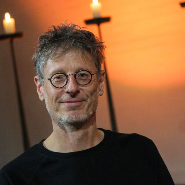 Eine männlich gelesene Person mit Brille und kurzen Haaren vor leuchtenden Kerzen.