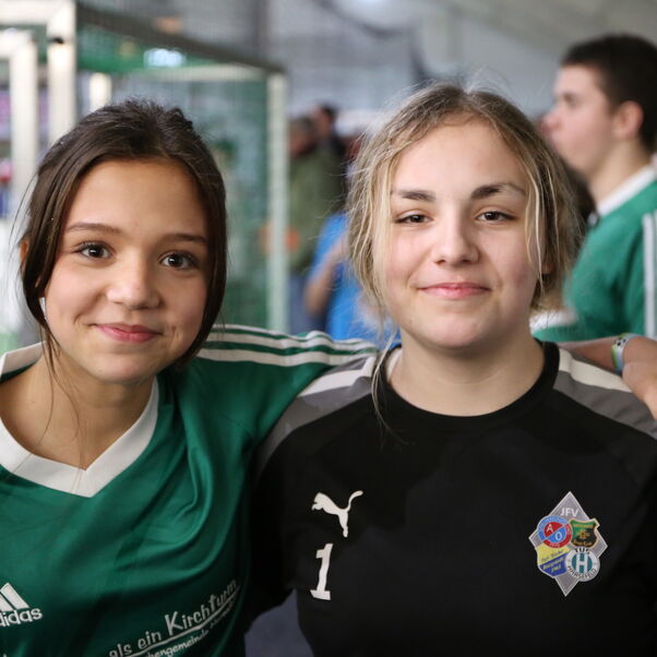 Zwei junge, weiblich gelesene Personen, eine legt den Arm um die andere, eine in grünem, eine in schwarzem Trikot.