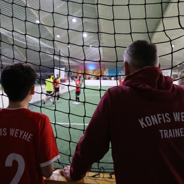 Zwei Personen blicken durch ein Netz auf einen Indoor-Fußballplatz.