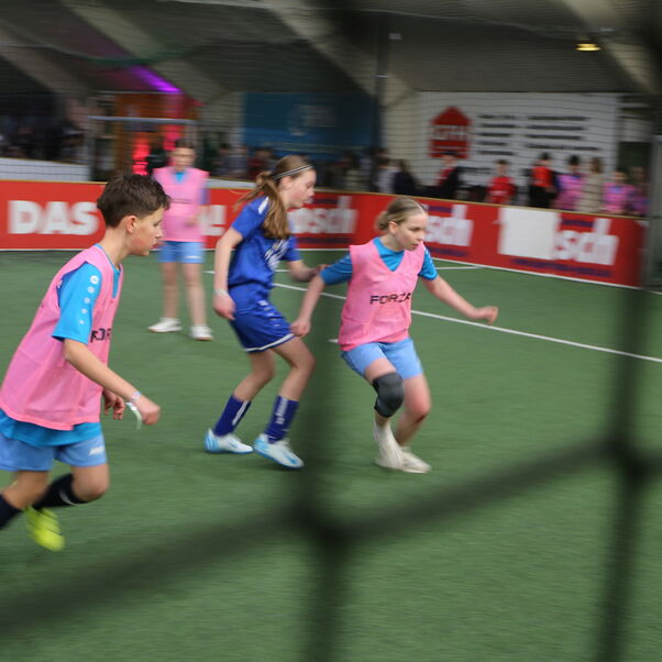 Blick durch ein Schutznetz auf ein Fußball-Spielfeld, auf dem junge Menschen in rosa und blauen Trikots spielen.
