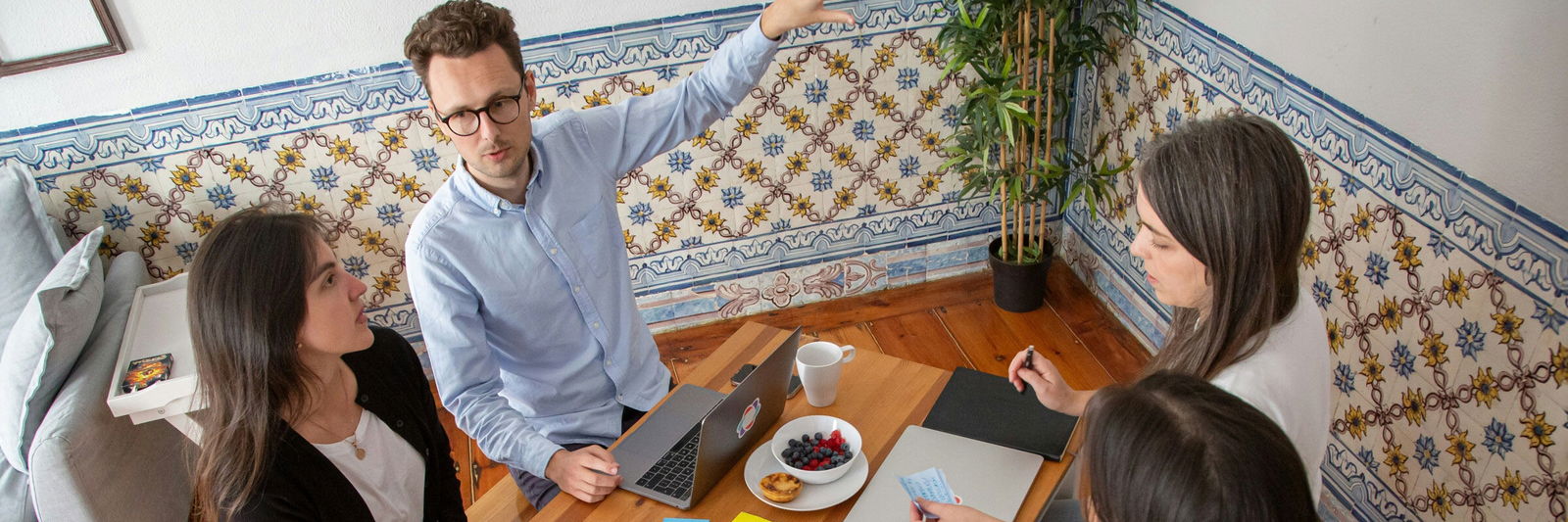 Eine Gruppe von fünf Menschen, die um einen Tisch sutzen. Notizbücher und Post-its. Ein Meeting-Raum mit Holzmöbeln, Sofa, Tapete und Grünpflanze. 