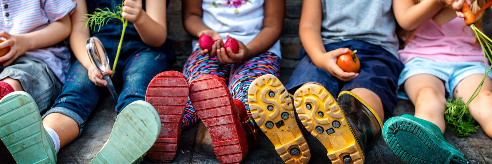 Eine Gruppe von Kindern sitzt auf einem Holzsteg und hält Gemüse in den Händen – ein Beispiel für die durch Kirchensteuer finanzierte Gemeinschaftsarbeit.