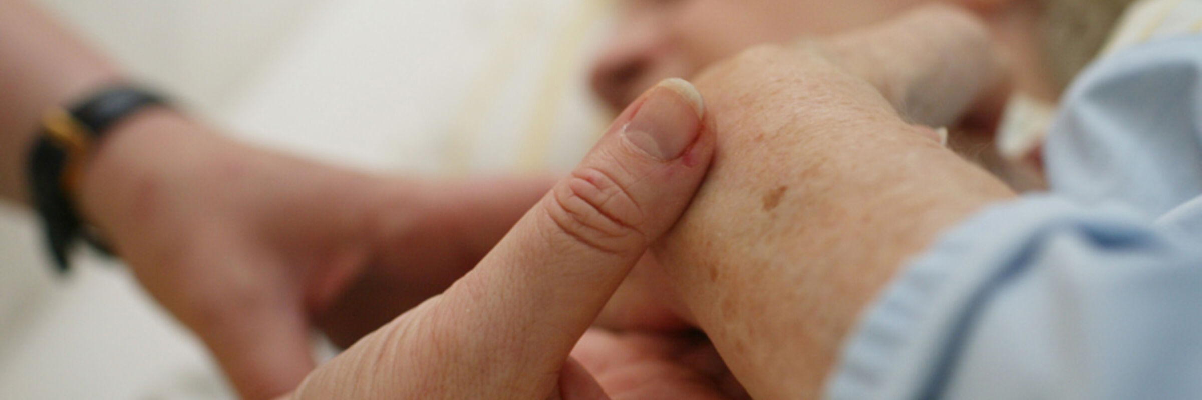 Das Bild zeigt im Vordergrund zwei Hände, die die Hand einer im Bett mit geschlossenen Augen liegenden Person hält. 