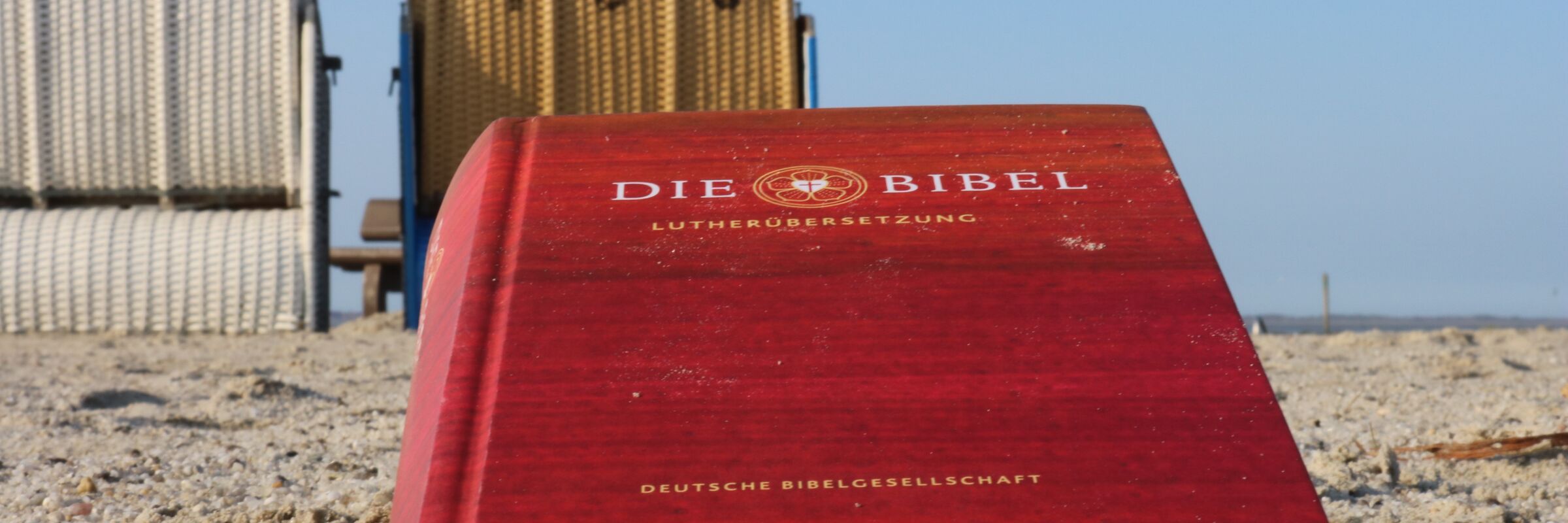 Eine Bibel im Sand am Strand. Im Hintergrund sind Strandkörbe zu erkennen. In der Urlaubsseelsorge kommt die Gute Nachricht Gottes natürlich auch an den Strand.