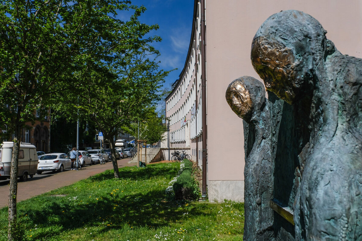 Das Bild zeigt zwei grau-goldene Skulpturen im Vordergrund vor einem rosafarbenen Gebäude an einer Straße mit belaubten Bäumen.