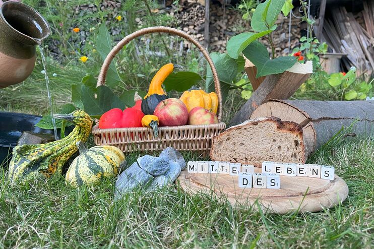 Ein Korb mit Obst und Gemüse, Kürbisse, ein Brot und ein Krug Wasser auf einer Wiese. Mit Scrabble-Würfeln sind die Worte Mittel des Lebens auf ein Brettgelegt.