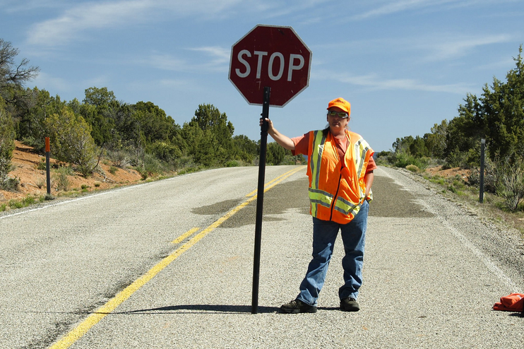 Ein Mann mit Bauarbeiter-Weste steht mit einem Stop-Schild auf einer Straße.
