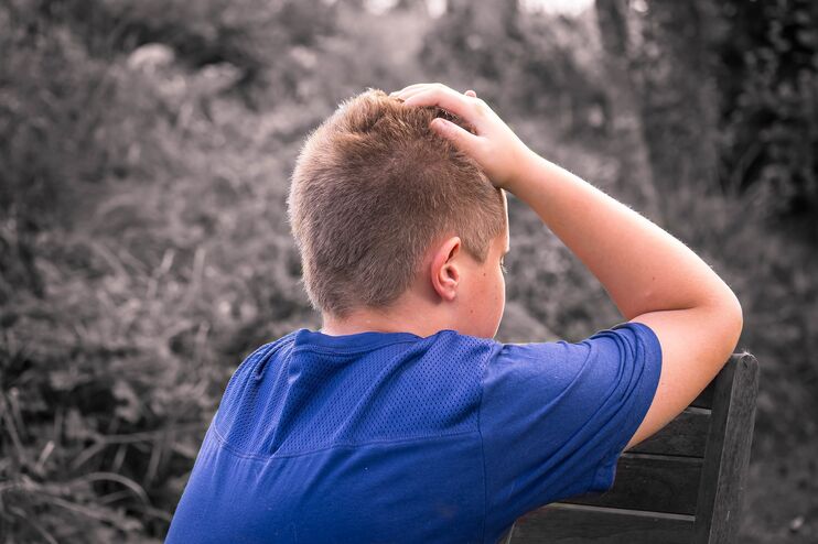Ein Junge im blauen T-Shirt stützt an einem Baum seinen Kopf in seine Hände.