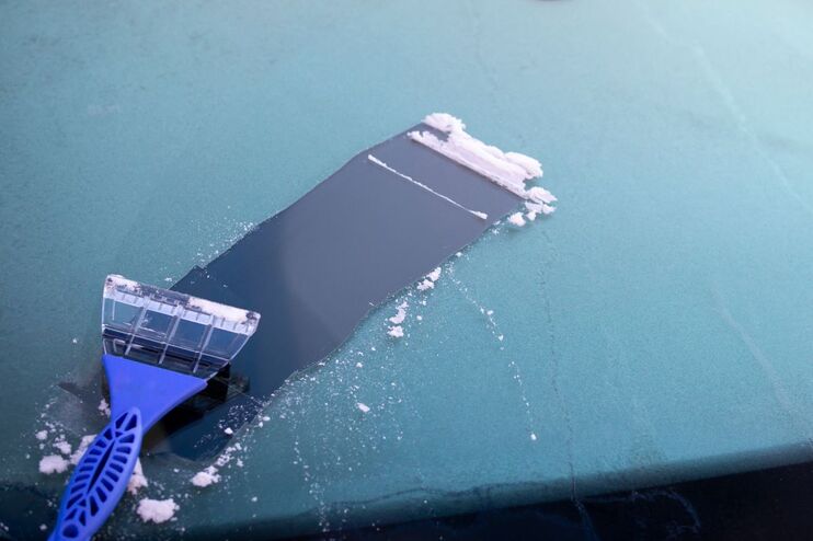 Ein Eiskratzer mit blauem Griff liegt auf einer vereisten Autoscheibe, auf der eine kleine Bahn freigeschoben wurde.