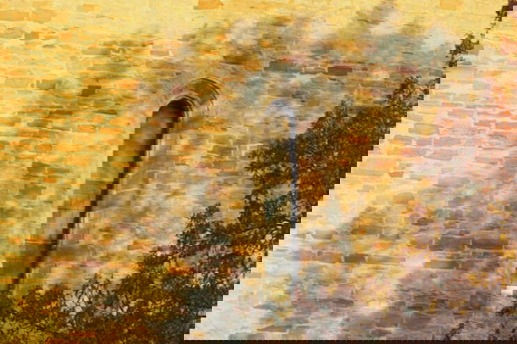 Eine Kirchenmauer mit einem kleinen Fenster. darüber liegt der Schatten eines Baumes, der vor der Kirche steht. Ein paar seiner Äste sind rechts im Bild noch zu sehen.