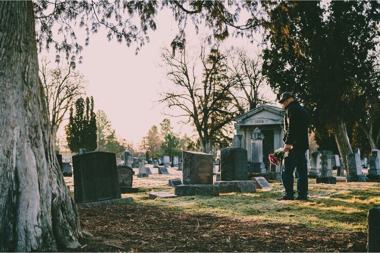 Ein Mann geht auf einem Friedhof zu einem Grab. Er hält einen Straß mit Blumen in der Hand. Sein Blick ist gesenkt.