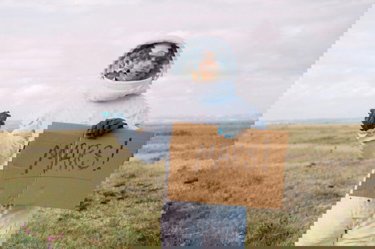 Eine als Mann lesbare Person in einem Raumanzug am Straßenrand und hält den Daumen raus. In der anderen Hand hält er ein Schild mit der Aufschrift MARS.