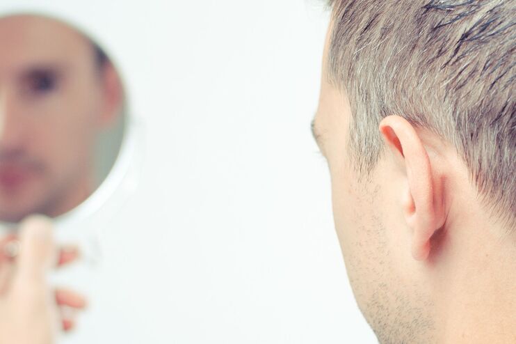Ein Mann betrachtet sich und seine grauen Haare kritisch in einem Handspiegel. 