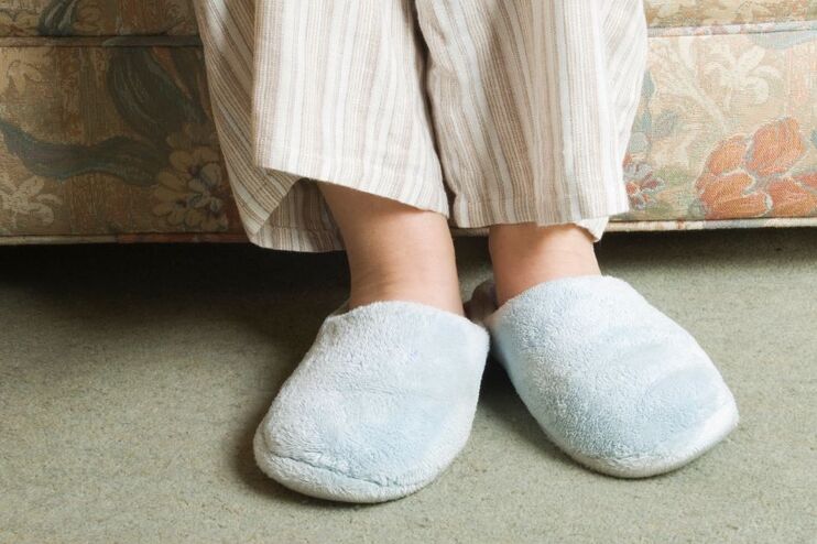 Nahaufnahme von Beinen, deren Füße in plüschigen Pantoffeln stecken, vor einem altmodischen Sofa.