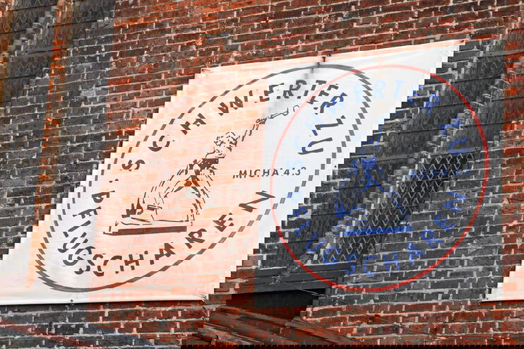 Ein weißes Transparent an einer Backsteinmauer mit der Aufschrift Schwerter zu Pflugscharen und der grafischen Darstellung eines Denkmals