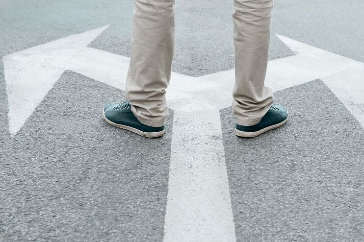 Eine Person mit grauer Hose steht auf einem weißen Pfeil auf der Straße. Der Pfeil teilt sich: nach links und rechts.