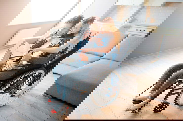 Eine als Frau lesbare Person umarmt von hinten eine als Mann lesbare Person im Rollstuhl, die am Laptop arbeitet.