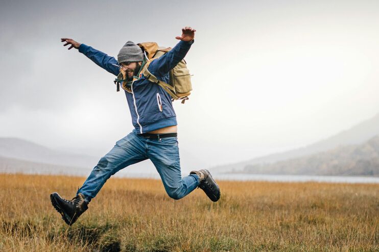 Ein Mann in Wandersachen und Rucksack springt auf einem Feld über einen Bachlauf.
