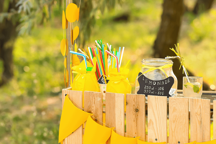 Auf einer grünen Wiese sieht man den Ausschnitt von einem Holz-Stand, an dem Zitronen- und Limetten-Limonade verkauft wird. Es gibt ein Preisschild. Der Stand ist mit gelben Wimpeln dekoriert.