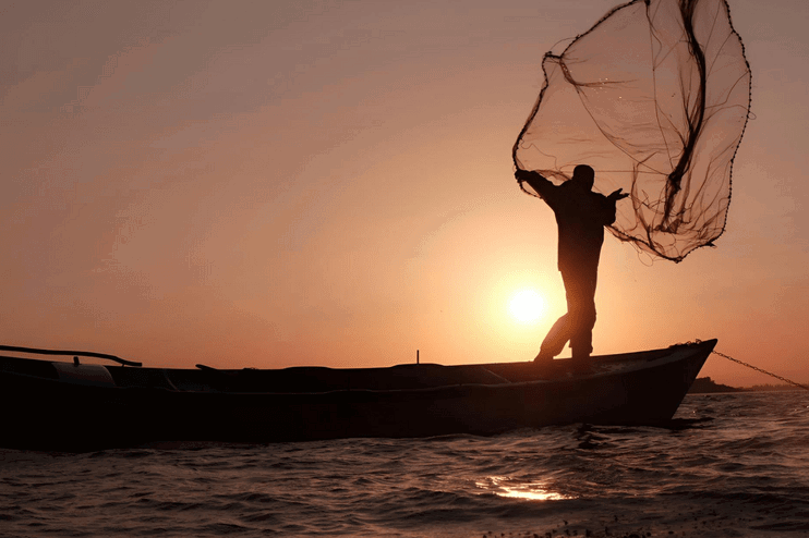 Ein Fischer wirft ein Netz von einem kleinen Boot aufs Meer vor einem Sonnenuntergang.