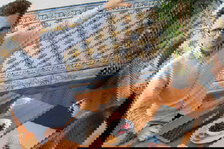 Eine Gruppe von fünf Menschen, die um einen Tisch sutzen. Notizbücher und Post-its. Ein Meeting-Raum mit Holzmöbeln, Sofa, Tapete und Grünpflanze. 