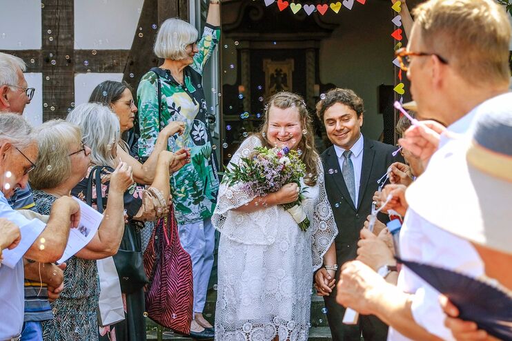 Vor einem Fachwerkhaus geht ein Hochzeitspaar durch eine Reihe freudiger Menschen, die Seifenblasen pusten.