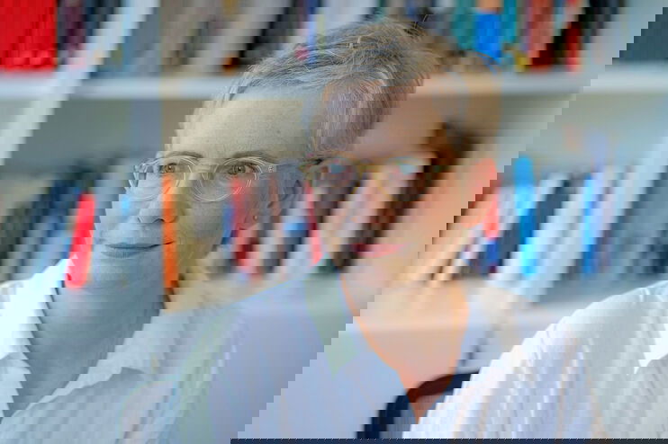 Der Bild zeigt eine weiblich lesbare Person mit kurzen hellblonden Haaren, einer hellen Brille, heller Hautfarbe und einer weißen Bluse. Die Person sitzt vor einem gefüllten Bücherregal.