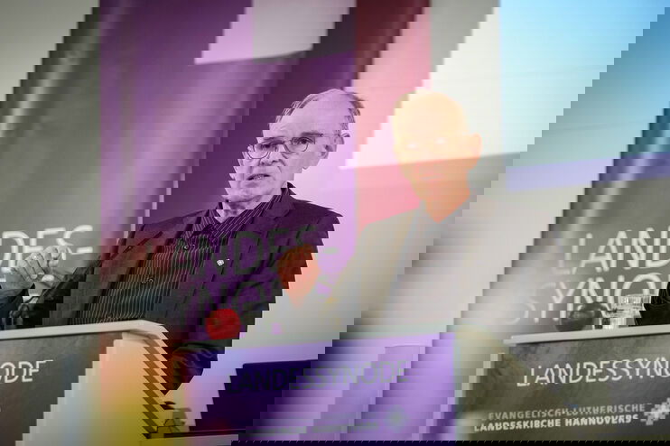 Eine männlich gelesene Person mit kurzen grauen Haaren und Brille hinter dem Redepult