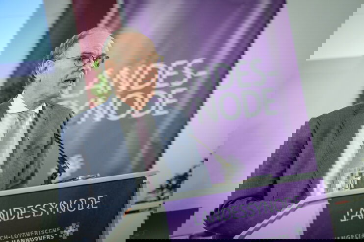 Eine männlich lesbare Person mit Brille und kurzen Haaren steht im Anzug an einem Pult und redet. 