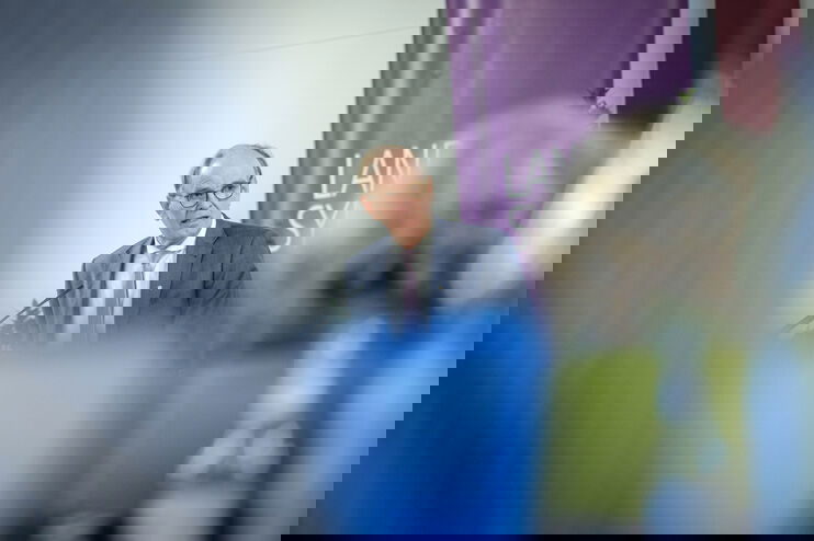 Eine männlich lesbare Person mit Brille und kurzen Haaren steht im Anzug an einem Pult und redet. Im Vordergrund sitzen Menschen.