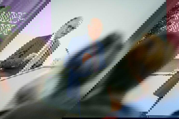 Eine männlich lesbare Person mit Brille und kurzen Haaren steht im Anzug an einem Pult und redet. 