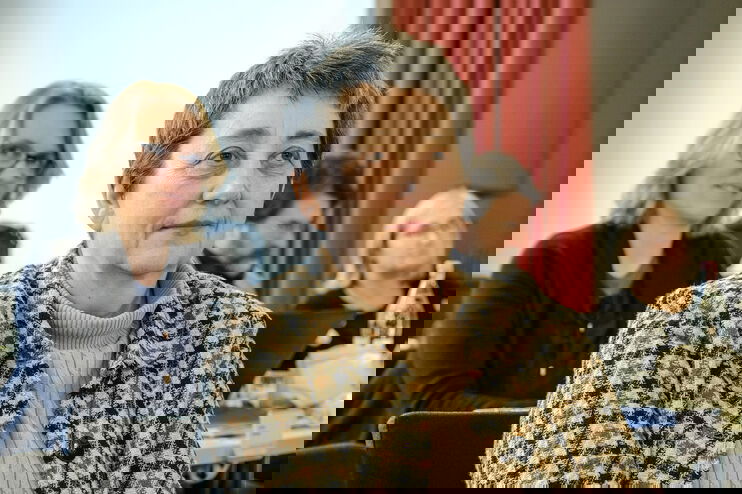 Eine weiblich lesbare Person mit karierter Bluse sitzt, mehrere weiblich lesbare Personen im Hintergrund