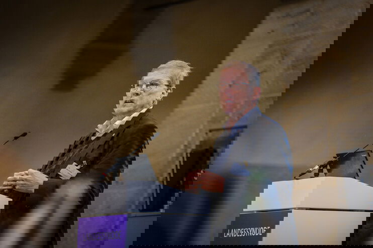 Eine männlich lesbare Person steht an einem Podium und spricht.