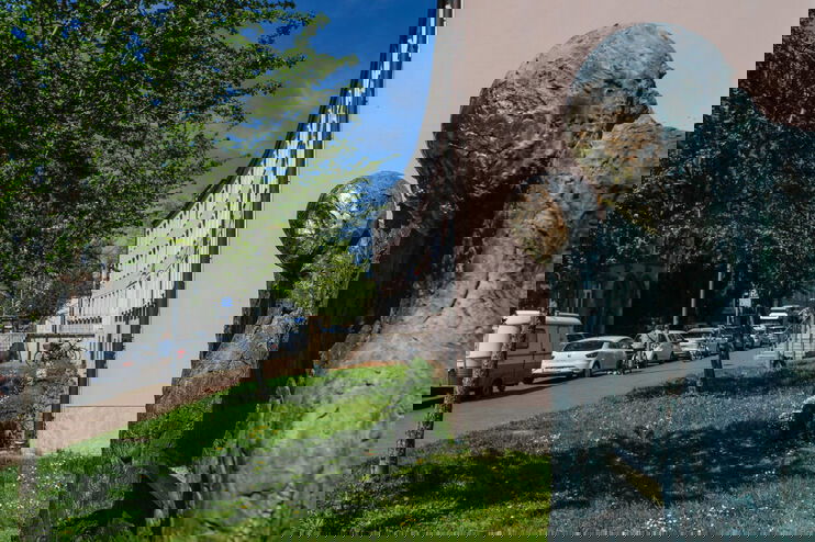 Das Bild zeigt zwei grau-goldene Skulpturen im Vordergrund vor einem rosafarbenen Gebäude an einer Straße mit belaubten Bäumen.