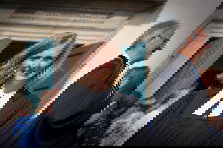 eine junge Pastorin steht vor einer Kirche. Sie hat blonde Haare, lächelt in die Kamera und trägt einen schwarzen Talar.