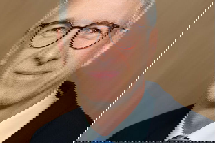 Ein Man lächelt in die Kamera. Er trägt einen schwarzen Anzug und eine blaue Krawatte. Der Mann hat graue, kurze Haare und trägt eine braune Hornbrille.