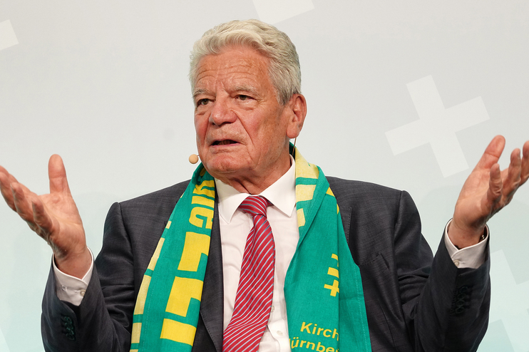 Ein älterer Mann mit grauem Haar und Kirchentagsschal spricht und hebt dabei beide Hände.