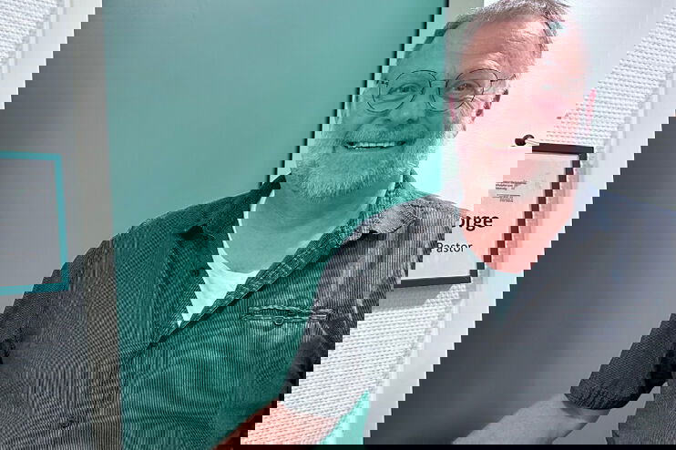 Ein Mann steht vor einer türkisen Tür. Er lächelt in die Kamera. Neben der Tür hängt ein Schild. Darauf steht: Schulseelsorger.