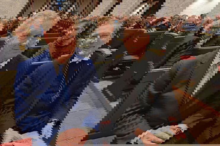 Zwei Männer, einer im Anzug, einer in Bischofskleidung, sitzen in der ersten Reihe einer Kirche.