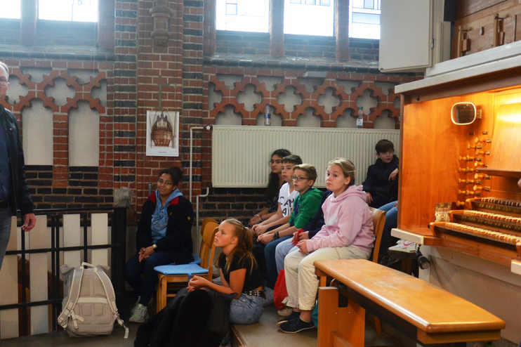 Mehrere Kinder sitzen neben einer Orgelbank und schauen einen schwarz gekleideten Mann an, der links im Bild steht.