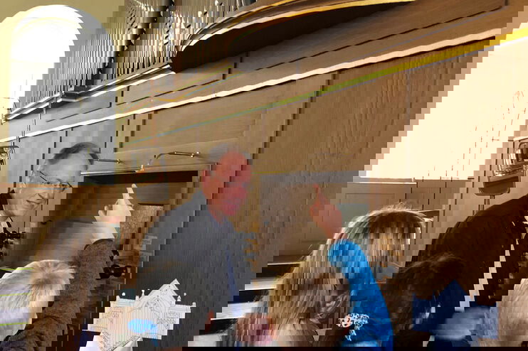 Ein Mann sitzt an der Orgel. Kinder stehen um ihn herum und stellen Fragen.