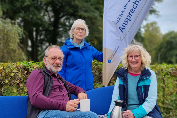 Ein Mann und eine Frau sitzen auf einer Bank mit blauen Sitzkissen. Hinter ihnen steht eine Frau in blauer Jacke. Im Hintergrund eine Hecke.