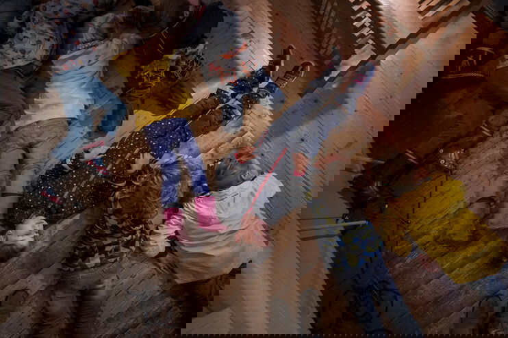 Blick von oben auf sechs Kinder, die auf einem Holzfußboden liegen, manche tragen Gummistiefel und Regenjacken. 