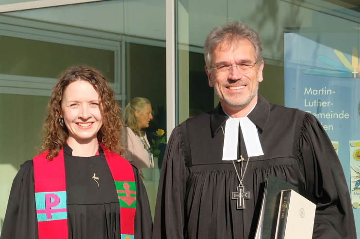 Eine junge Frau steht neben einem älteren Mann, beide tragen schwarze Talare. Die Frau trägt darüber eine bunte Stola. 