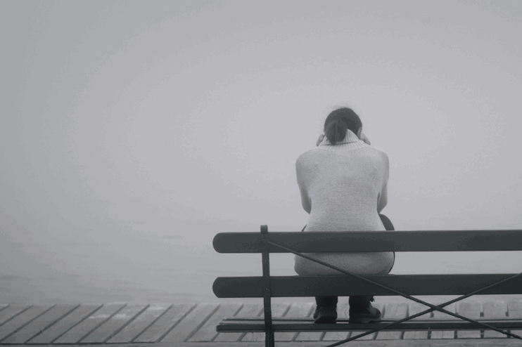 Eine junge Person hockt im Nebel auf einer Bank, alles ist grau.