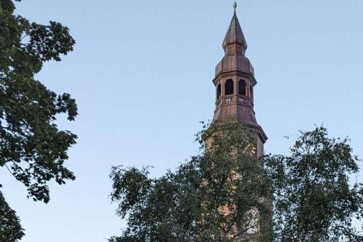Über grüne Baumwipfel ragt ein Kirchturm in den blauen Himmel.